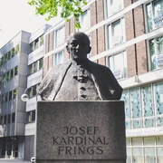 Joseph Frings Monument Das Joseph-Frings-Denkmal zeigt eine bronzene Büste des Kardinals auf einem steinernen Sockel, umgeben von modernen Gebäudeansichten.The Joseph Frings monument shows a bronze bust of the cardinal on a stone plinth, surrounded by modern views of buildings.