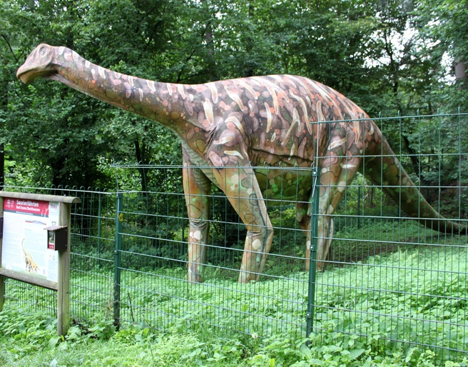 Modell eines Megalosauriers an den Saurierfährten in Bad Essen – Barkhausen. Dinosauriermodell in einem grünen Park hinter einem Metallzaun, neben einem Infotafel.