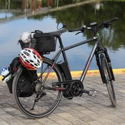 Ein schwarzes Fahrrad mit Gepäckträger und Helm steht auf gepflastertem Boden vor ruhigem Wasser.