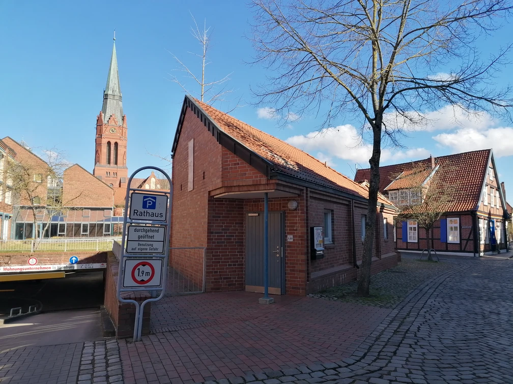 Toiletten Rathaus Tiefgarage Rote Backstein-Toilettenanlage in der Nähe des Rathauses, mit Zugang zur Tiefgarage und Kirchturm im Hintergrund.