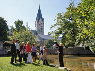 Eine Gästeführerin erklärt einer Gruppe die Paderquellen in Paderborn, im Hintergrund ein Kirchturm.