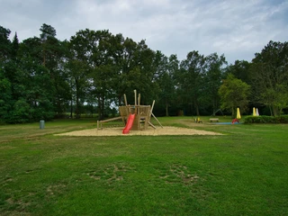 Spielplatz Offener Spielplatz auf einer Wiese, im Zentrum ein hölzernes Klettergerüst mit roter Rutsche, umgeben von Bäumen.