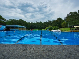 Schwimmbecken Ein glitzerndes, blaues Schwimmbecken im Freien, umgeben von Bäumen und Liegestühlen am Beckenrand.