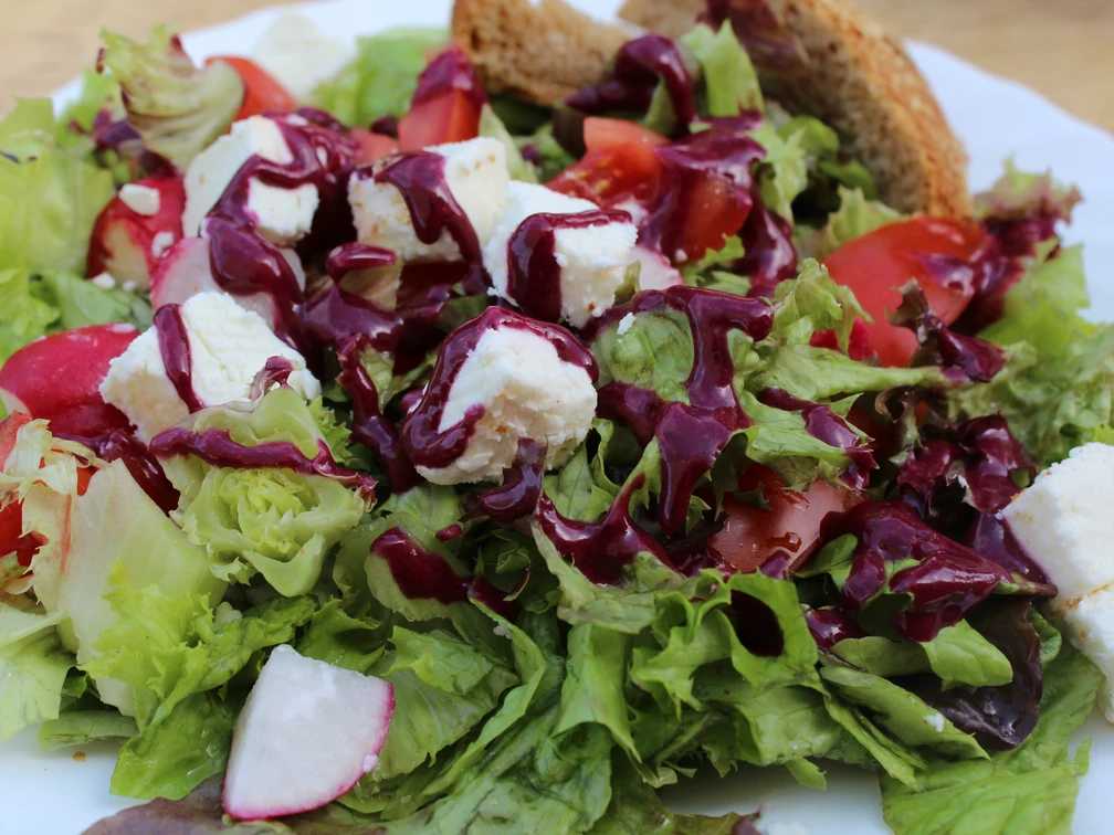 Ein frischer, bunter Salat mit Feta-Würfeln, Radieschen, Tomaten und rotem Dressing auf einem weißen Teller.