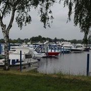 Wieltsee Boote liegen vertäut an Stegen eines ruhigen Sees, umrahmt von grünen Bäumen und bewölktem Himmel.