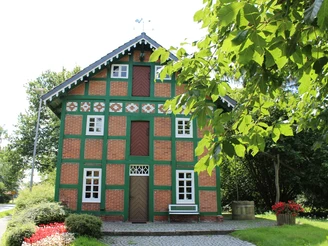 Weyhe Spieker Lahausen Rotes Fachwerkhaus mit grünem Gebälk, umgeben von Bäumen, mit Bank und Blumen vorne rechts.