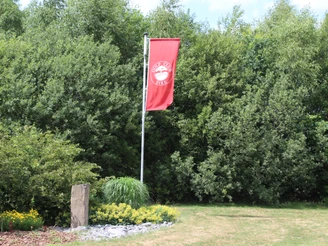 Syke Golfplatz Eine rote Fahne des Golfclubs Syke weht, umgeben von grünem Buschwerk und gepflegtem Rasen.