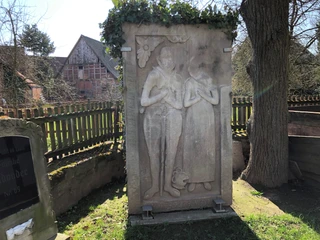 Kapelle Röhden Schlüsselburg Zwei antike Reliefs aus Stein, unter Efeuranken befestigt, stehen neben einem Baum mit Holzzaun.