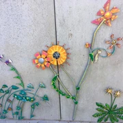 Blumenvielfalt am Lieblingsort Tange Eine Wand ziert ein farbenfrohes Kunstwerk mit fantasievoll gestalteten Metallblumen. Die Blüten strahlen in leuchtenden Orange- und Gelbtönen, ergänzt durch stilisierte grüne Blätter. Es verleiht der Wand einen lebhaften und kreativen Ausdruck.
