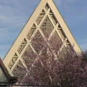 Kirche Eine Kirche mit modernem dreieckigem Dach und geometrischen Mustern, umgeben von blühenden Bäumen.