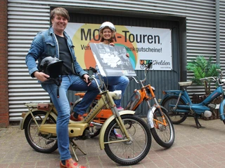 Mofa-Helden Zwei Personen lächeln auf Mofas vor einer Werbetafel für Mofa-Touren, ausgestattet mit Helmen.