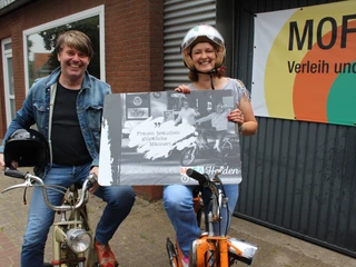 Mofa-Helden Zwei Personen auf alten Mofas lächeln, die Frau hält ein humorvolles Plakat in der Hand.