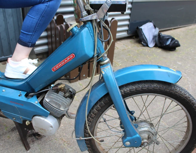 Mofa-Helden Ein blauer Zündapp-Mofa mit Reifenprofil, auf dem eine Person mit weißen Sneakern sitzt.