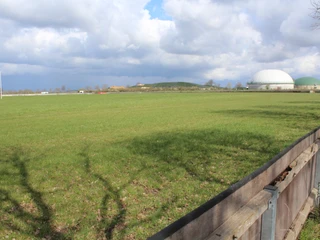 Rennbahn Schwarme Grüne Wiese mit einem Holzzaun im Vordergrund; im Hintergrund zwei große Biogasanlagenkuppeln.