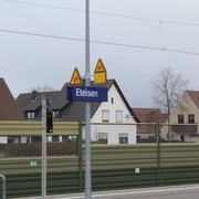 Bahnhof Etelsen Bahnhofsschild "Etelsen" vor Wohngebäuden, im Hintergrund teils bewölkter Himmel und Oberleitungen.