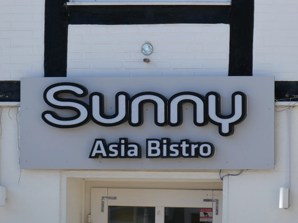 Sunny Asia Bistro Leuchtendes Schild mit der Aufschrift "Sunny Asia Bistro" an einer Gebäudefassade mit Fachwerkelementen.