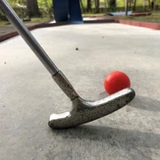 Ein silberner Minigolfschläger im Holzstil trifft auf einen roten Ball auf einer Betonanlage.