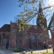 Backsteinkirche mit hohem Turm und Spitzbogenfenstern, umrahmt von Bäumen unter klarem, blauen Himmel.