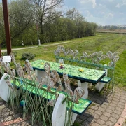 Lieblingsort am Wanderweg Das Bild zeigt einen dekorativen Rastplatz entlang eines Wanderwegs. Es umfasst kunstvoll gestaltete Tische und Bänke, verziert mit metallischen Pflanzenmotiven, die an Blätter und Gräser erinnern. Im Hintergrund erstreckt sich eine grüne Landschaft unter einem leicht bewölkten Himmel.