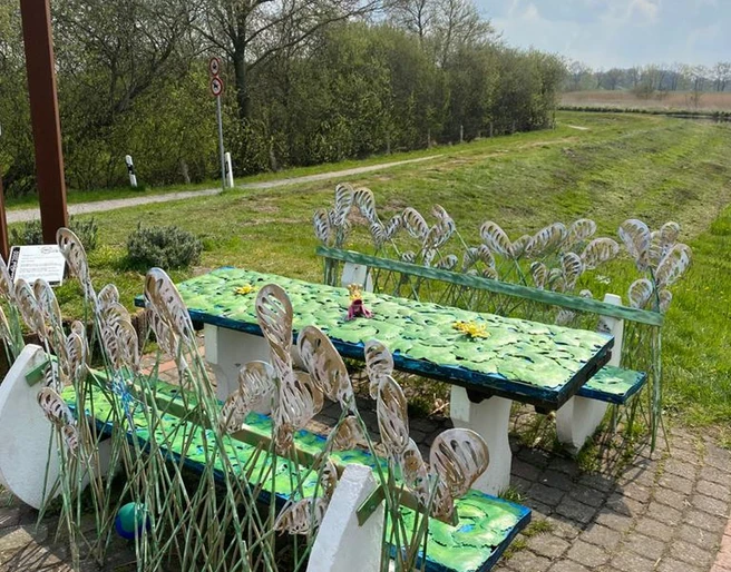 Lieblingsort am Wanderweg Das Bild zeigt einen dekorativen Rastplatz entlang eines Wanderwegs. Es umfasst kunstvoll gestaltete Tische und Bänke, verziert mit metallischen Pflanzenmotiven, die an Blätter und Gräser erinnern. Im Hintergrund erstreckt sich eine grüne Landschaft unter einem leicht bewölkten Himmel.