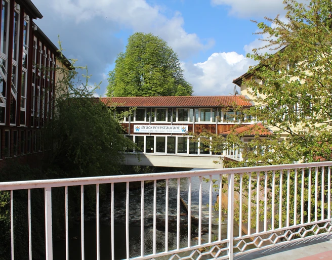 Brückenrestaurant Ein Restaurant auf einer Fußgängerbrücke verbindet zwei Gebäude über einem Fluss in üppiger Natur.