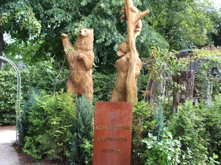 KreativWerkstatt am Wall Holzskulpturen von Bären, umgeben von grünem Laub und niedrigen Büschen, im Freien platziert.