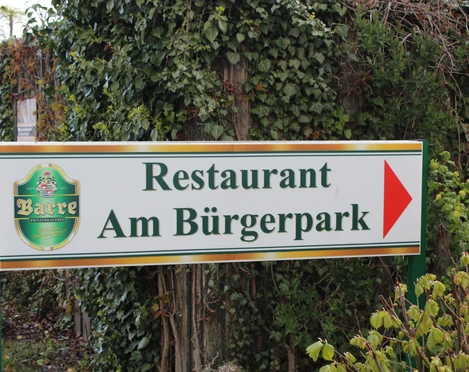 Restaurant am Bürgerpark Schild des Restaurants Am Bürgerpark mit Logo, umgeben von üppigem Grün und blühenden Pflanzen.