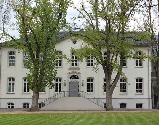 Besselsche Hof Ein weißes, zweistöckiges Gebäude mit symmetrischen Fenstern, umgeben von grünen Bäumen und Rasenflächen.