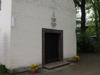 Eingang der Kapelle Nordel mit dunkelbrauner Tür, gerahmt von blühenden Pflanzen in Töpfen.