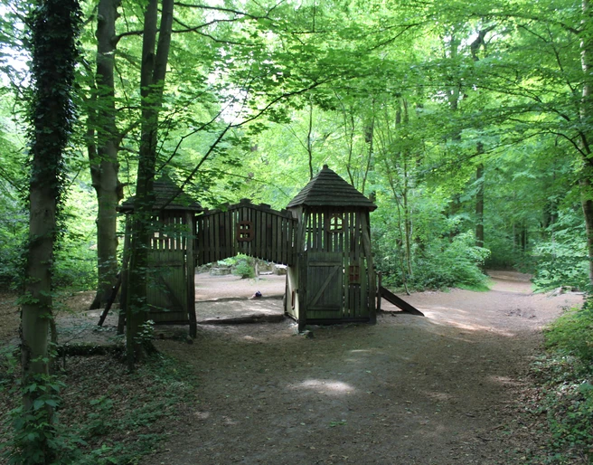 Waldspielplatz Heiligenberg Ein naturnaher Spielplatz im Wald mit einer Holzburg und umgebenden hohen Bäumen unter grünem Blätterdach.
