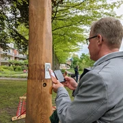 Hörtstation 02 Rheine-Bürgermeister Dr. Lüttmann Ein Mann scannt einen QR-Code an einer hölzernen Hörstation in einem parkähnlichen Umfeld.