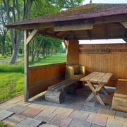 Rastplatz in Klauhörn Der Rastplatz in Klauhörn zeigt einen überdachten Unterstand mit Holztisch und Bänken auf gepflastertem Boden. Im Hintergrund sind grüne Wiesen und hohe Bäume zu sehen, die eine malerische Landschaft bilden.