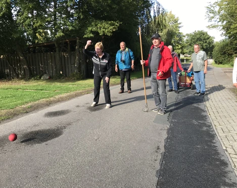 Boßelspaß Auf einer ruhigen Landstraße wird das traditionelle Boßeln gespielt. Eine Gruppe von Menschen, darunter eine Frau im Vordergrund, konzentriert sich auf das Werfen einer roten Boßelkugel über die Asphaltstraße, begleitet von lächelnden Teilnehmern und einem Bollerwagen voller Zubehör im Hintergrund. Die umgebende grüne Landschaft schafft eine friedliche Kulisse an diesem sonnigen Tag.