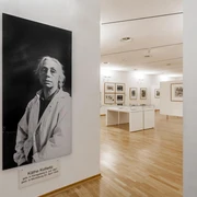 Käthe Kollwitz Museum Cologne Das Bild zeigt einen hellen Raum im Käthe Kollwitz Museum in Köln. Links ist ein großes Schwarz-Weiß-Porträt von Käthe Kollwitz in einem weißen Kittel vor einer Wand zu sehen. Im Hintergrund befinden sich Ausstellungsstücke in Vitrinen und an Wänden auf einem Parkettboden.The picture shows a bright room in the Käthe Kollwitz Museum in Cologne. On the left is a large black and white portrait of Käthe Kollwitz in a white coat in front of a wall. In the background are exhibits in display cases and on walls on a parquet floor.
