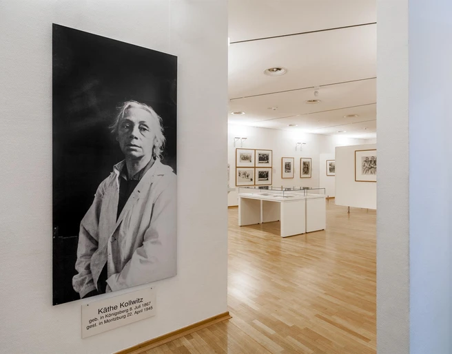 Käthe Kollwitz Museum Cologne Das Bild zeigt einen hellen Raum im Käthe Kollwitz Museum in Köln. Links ist ein großes Schwarz-Weiß-Porträt von Käthe Kollwitz in einem weißen Kittel vor einer Wand zu sehen. Im Hintergrund befinden sich Ausstellungsstücke in Vitrinen und an Wänden auf einem Parkettboden.The picture shows a bright room in the Käthe Kollwitz Museum in Cologne. On the left is a large black and white portrait of Käthe Kollwitz in a white coat in front of a wall. In the background are exhibits in display cases and on walls on a parquet floor.
