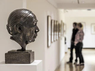 Käthe Kollwitz Museum Cologne Eine Bronzebüste der Künstlerin Käthe Kollwitz steht seitlich auf einem Podest, während im Hintergrund zwei Personen in einer Galerie die Kunstwerke betrachten.A bronze bust of a woman's profile is displayed in the foreground, with visitors observing artworks in a lit hallway in the background at the Käthe Kollwitz Museum in Cologne.