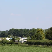 Campingplatz mit Wohnwagen zwischen grünen Büschen und Bäumen, entferntes Waldstück im Hintergrund.