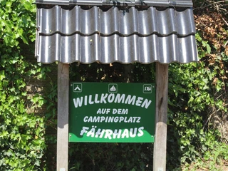 Campingplatz Fährhaus Grünes Schild mit weißer Aufschrift "Willkommen auf dem Campingplatz Fährhaus", umrahmt von Pflanzen.