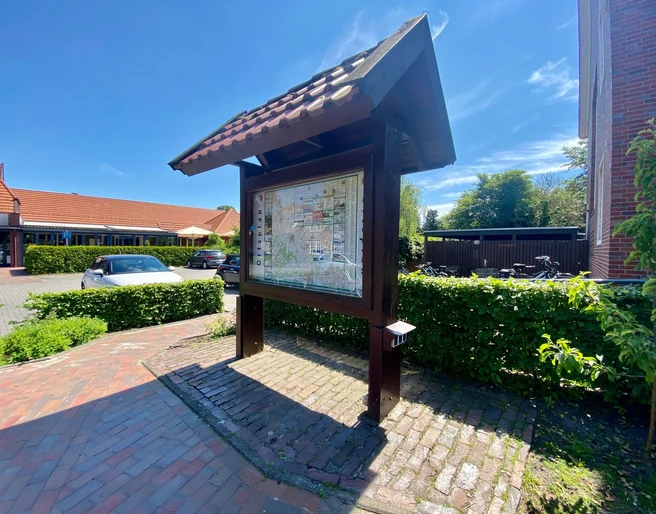 Ladestation E-Bike.jpg Karte mit Orten unter einem Dach aus Holz, im Sonnenlicht, auf Backsteinweg.