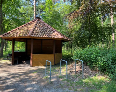 Rastplatz Mansholter Holz.jpg Ein kleiner Rastplatz mit einem hölzernen Pavillon und roten Ziegeln, umgeben von dichtem Grün.