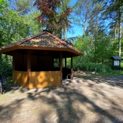 Rastplatz Mansholter Holz2.jpg Ein hölzerner Pavillon mit schattenspendendem Dach steht an einem Waldweg, umgeben von grünen Bäumen und einem Fahrradständer.