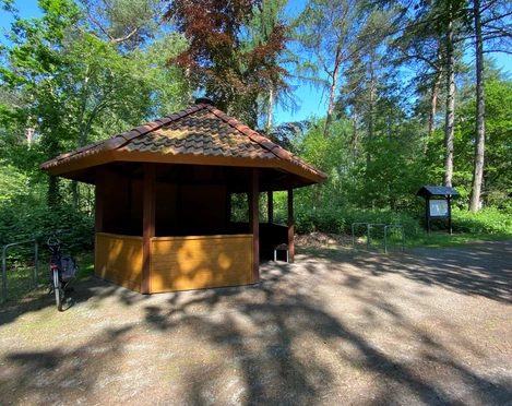 Rastplatz Mansholter Holz2.jpg Ein hölzerner Pavillon mit schattenspendendem Dach steht an einem Waldweg, umgeben von grünen Bäumen und einem Fahrradständer.