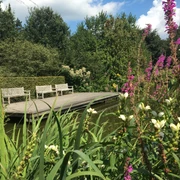 IMG_5367.JPG Malerische hölzerne Brücke über einen ruhigen Fluss, umrahmt von üppiger Natur und bunten Blumen.