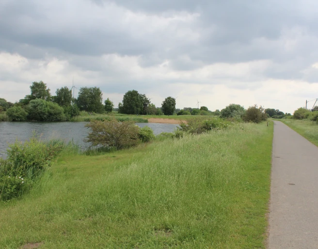 Alte Weser Flussufer mit Fuß- und Radweg, umgeben von saftigem Grün; im Hintergrund Windmühlen unter bewölktem Himmel.