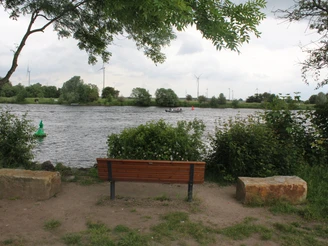 Reisegarten Weye Eine Holzbank mit zwei Steinen am Weserrand, umgeben von grüner Vegetation und Blick auf Windräder.