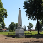 Denkmal auf einem Marktplatz, umrahmt von Bäumen, mit einem schwarzen Zaun davor. Wiese im Hintergrund.