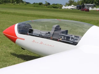 Ein weißes Segelflugzeug, rot markiert, steht auf einer grünen Wiese, bereit zum Starten.