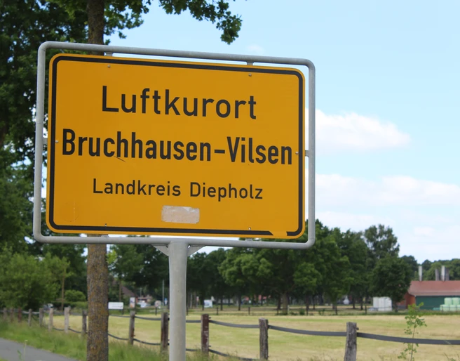 Ortsschild Ortsschild für den Luftkurort Bruchhausen-Vilsen im Landkreis Diepholz, umgeben von grünen Bäumen.