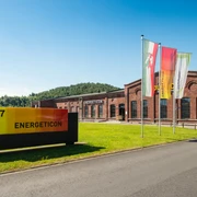 Eingangsbereich zu einem industriell aussehenden Gebäude mit Fahnen und einer Wiese davor.