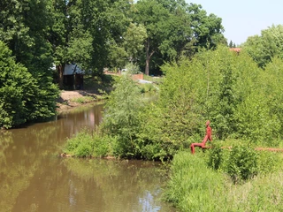 Skulptur "Der rote Mann" Eine rote Skulptur eines sitzenden Mannes am Ufer eines Flusses, umgeben von üppigen Bäumen.